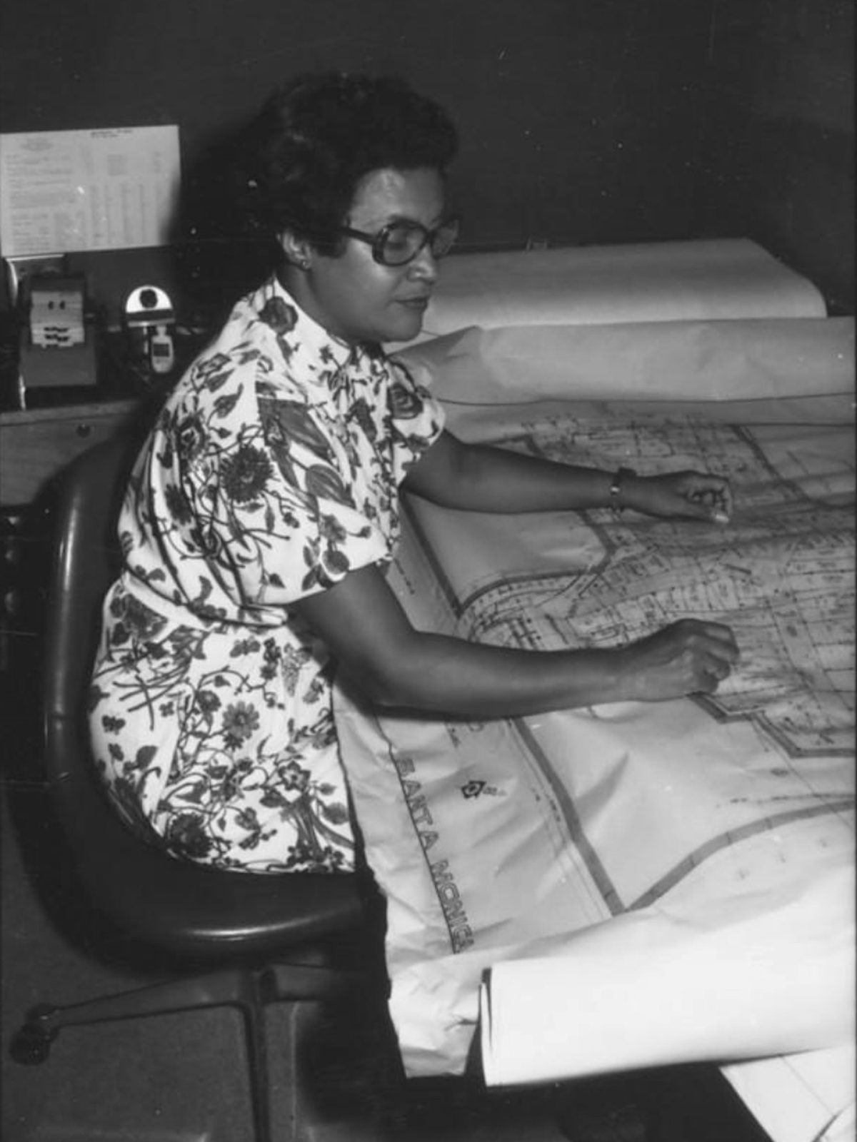 a woman reviewing a large architectural plan at her desk, 1970s