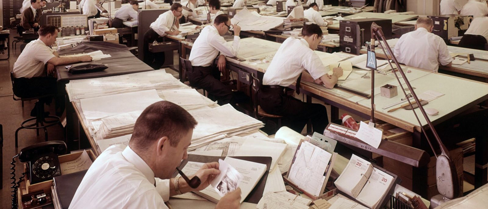 rows of draftsmen at drawing tables in a large drafting room, 1960s