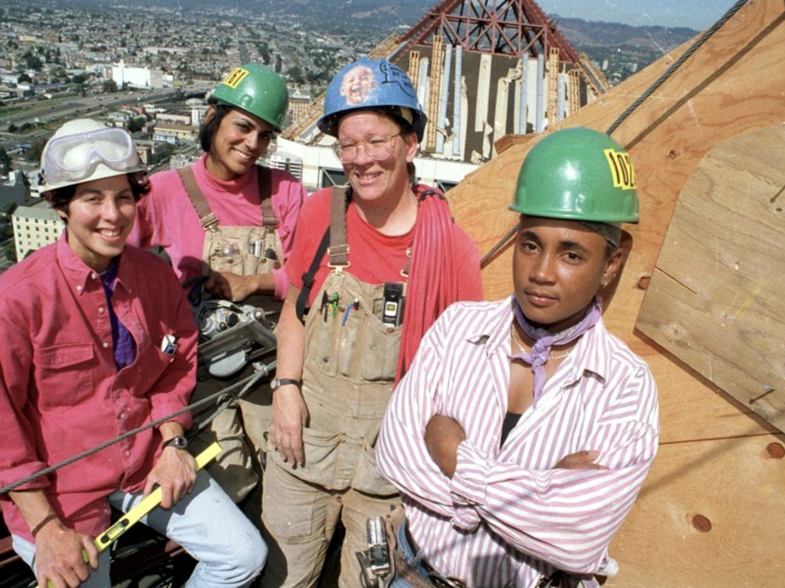 four tradeswomen in hard hats on a rooftop job site above a city