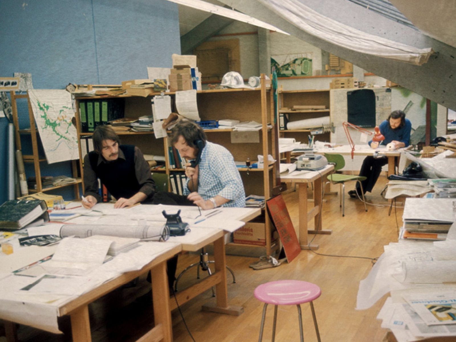 architects at work in a cluttered studio full of plans and models, 1970s