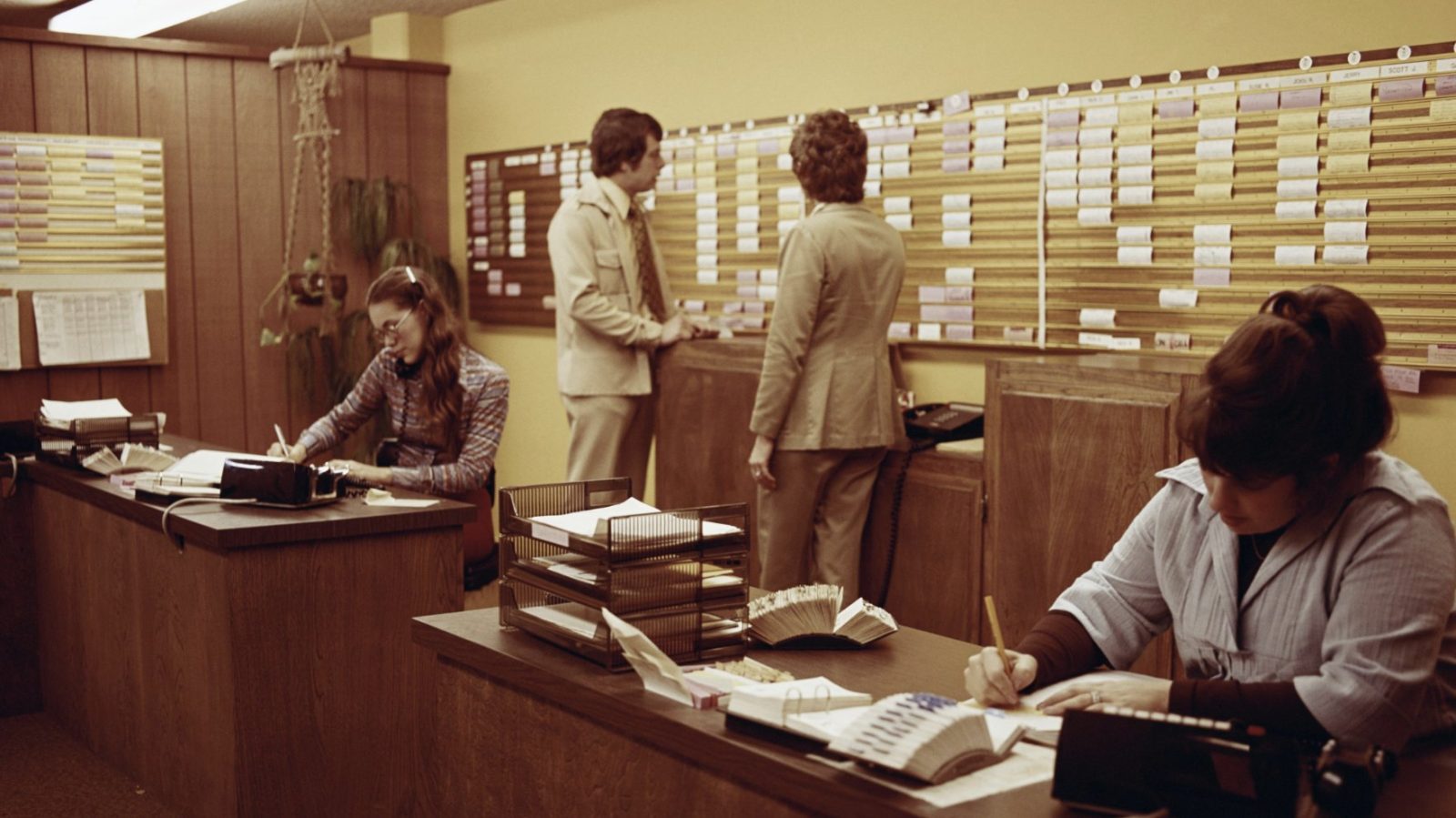 office staff working at desks in front of a wall-length scheduling board, 1970s
