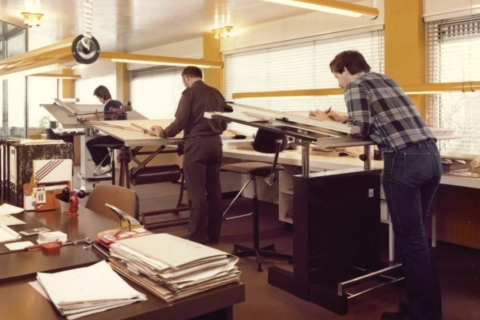drafters at standing drawing boards and a man at a desk in a bright 1970s office