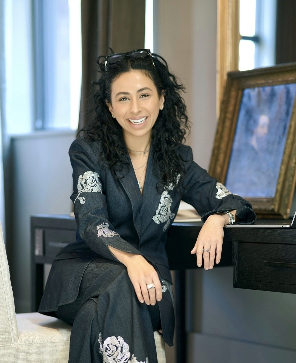 hailey george in a dark denim suit with silver embroidered roses, smiling, seated at a desk