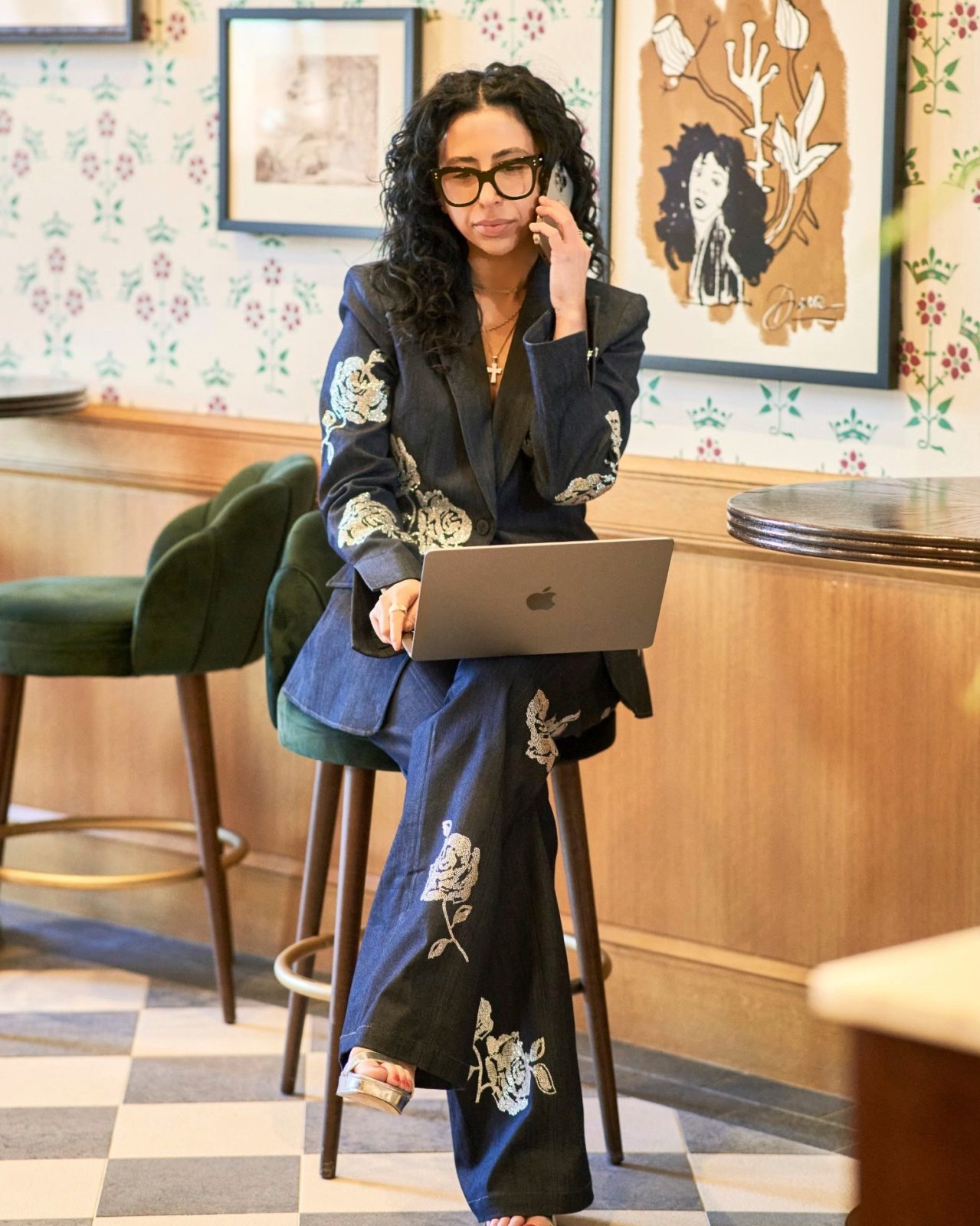 hailey george in an embroidered denim suit and glasses, on the phone with a laptop on her lap, seated on a green velvet stool