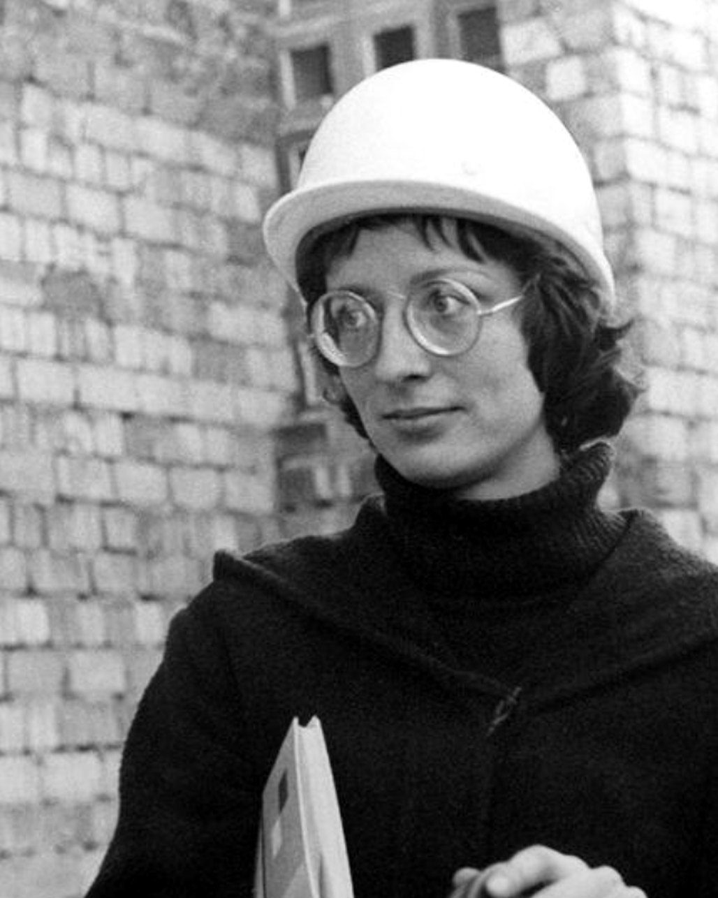 a woman in a hard hat and round glasses on a building site, black and white