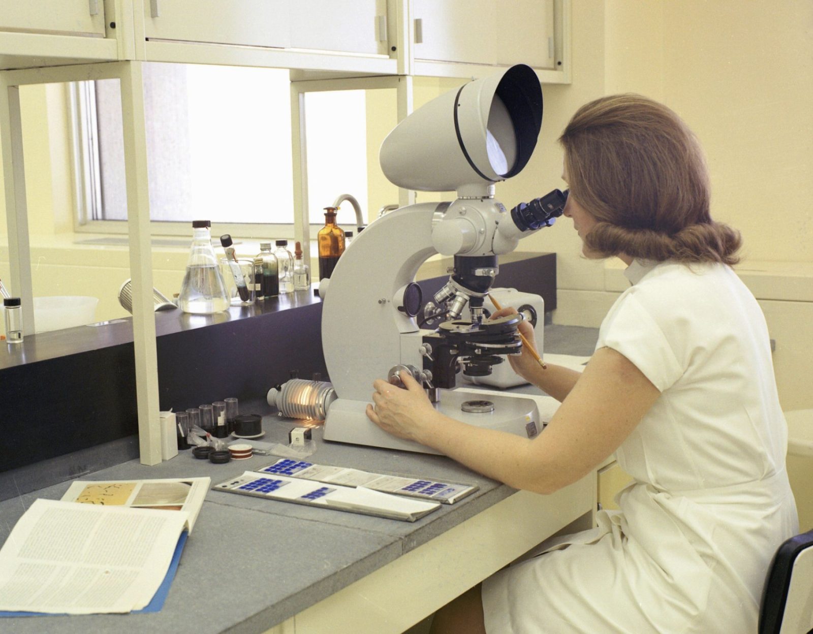 a lab technician looking into a microscope at a laboratory bench, 1960s