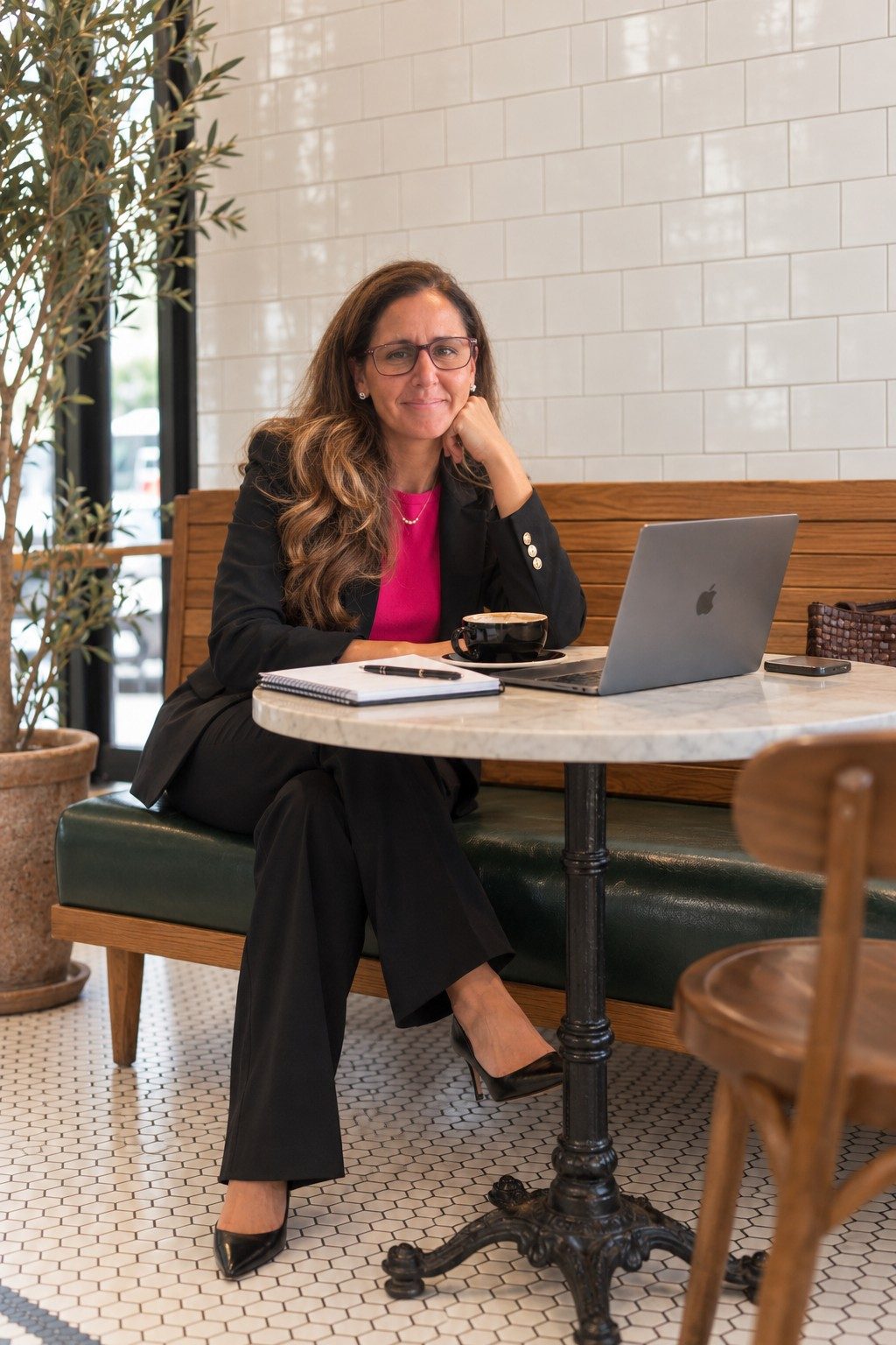 michelle danubio in a black suit and pink top, seated at a marble cafe table with a laptop and coffee