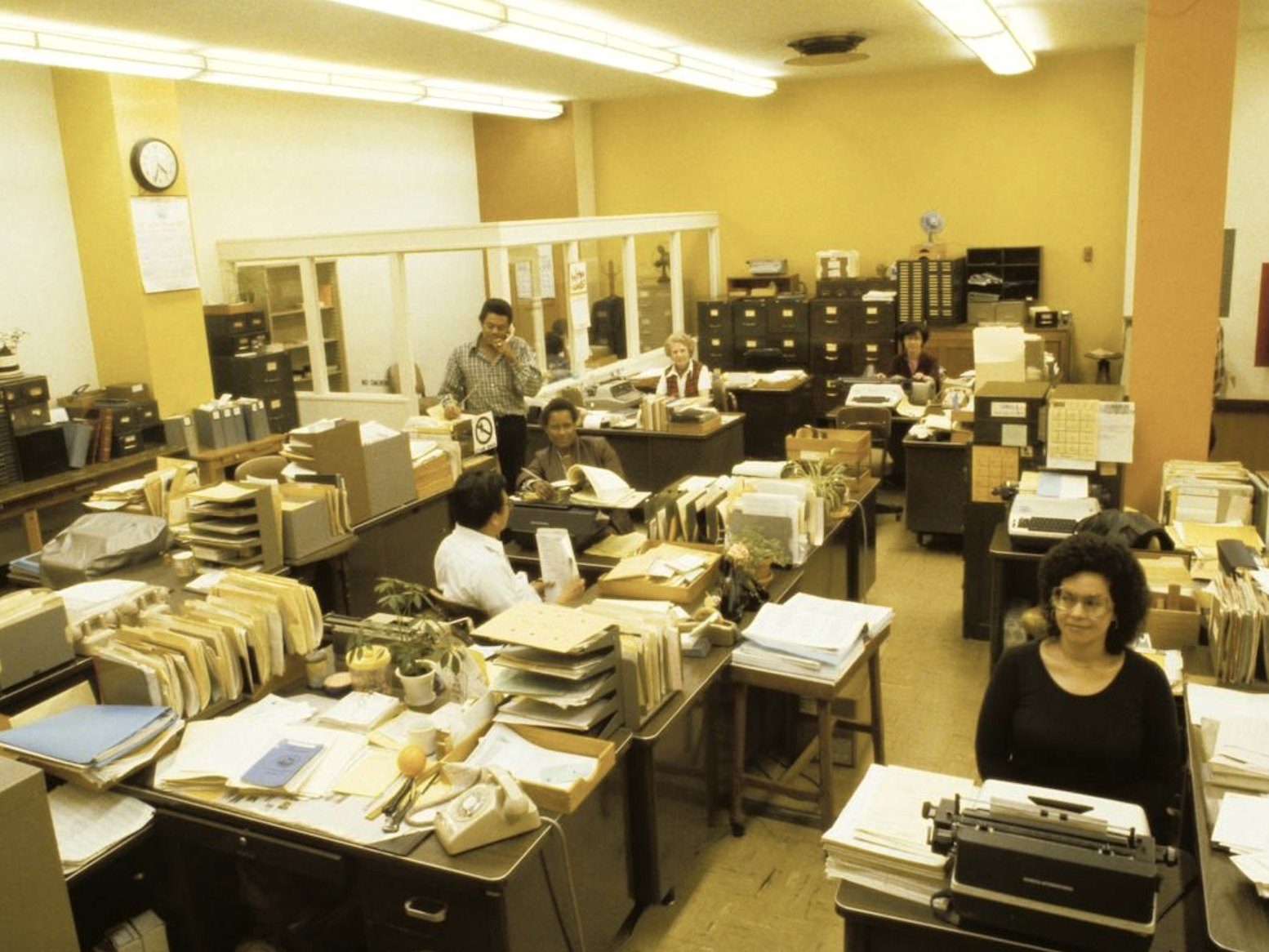 staff at desks stacked with files and typewriters in a yellow-walled office at muni’s presidio division, san francisco, late 1970s