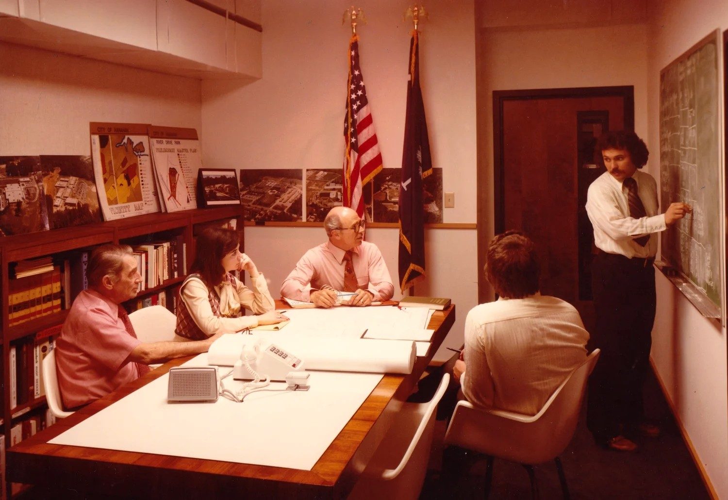 a project team meeting around a conference table while a colleague sketches at a chalkboard, with site plans and aerial photos on the shelves, 1970s